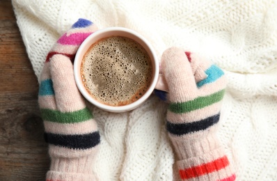 Woman with knitted mittens holding hot winter drink on warm plaid, top view. Cozy season