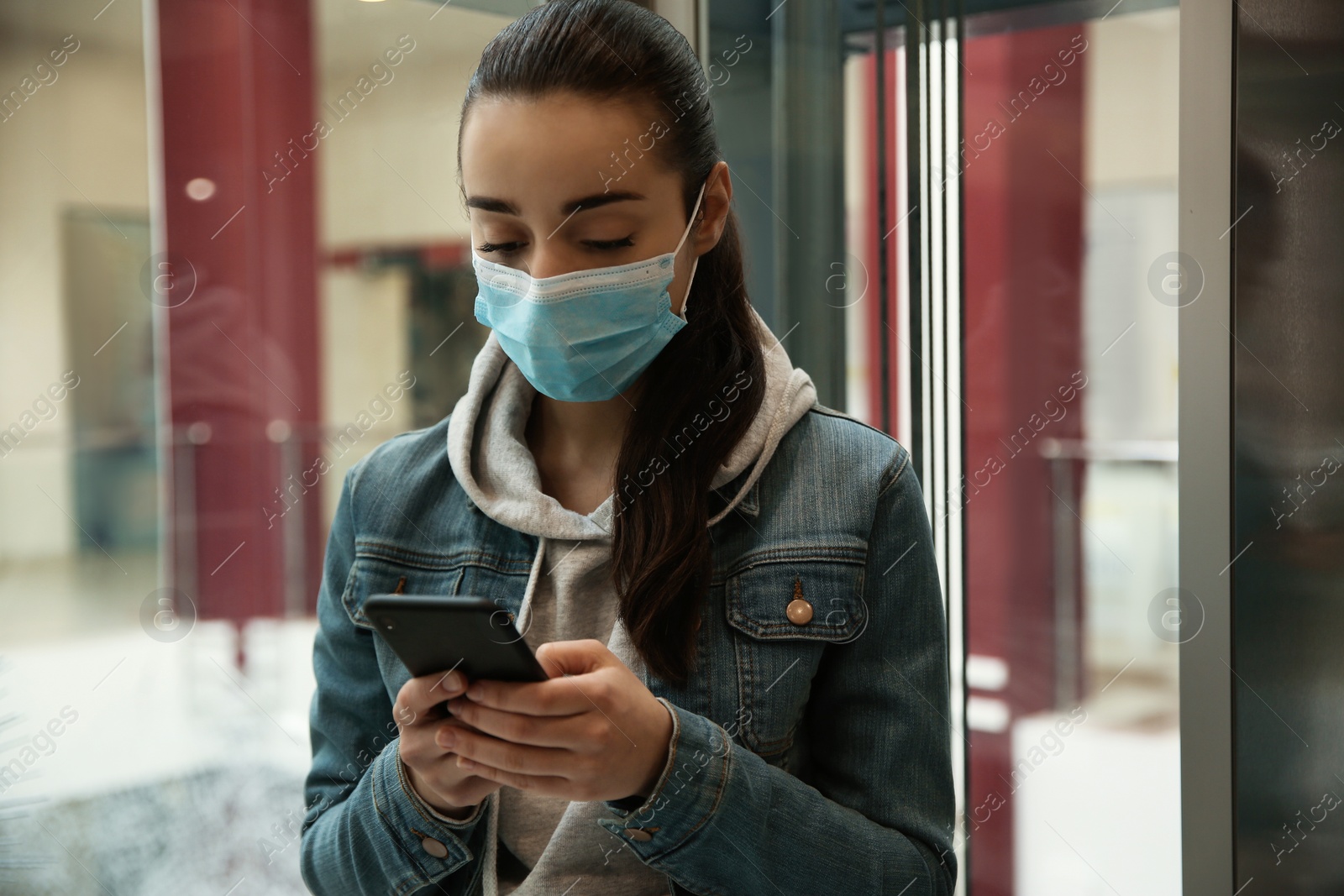 Photo of Young woman with disposable mask and smartphone in elevator. Dangerous virus
