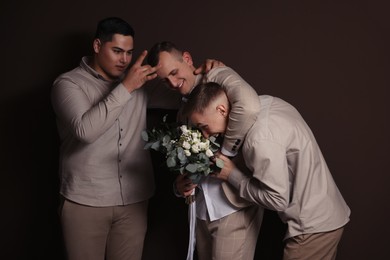 Happy groom with bouquet and his groomsmen having fun on brown background