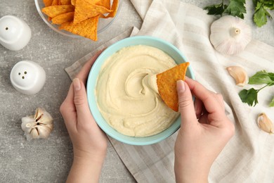 Woman dipping nacho chip into hummus at grey table, top view