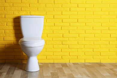 Photo of New clean toilet bowl near yellow brick wall indoors, space for text. Interior design