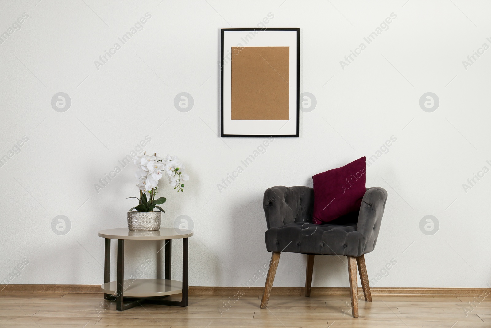 Photo of Comfortable armchair, cushion, table with orchid and picture frame indoors. Interior design