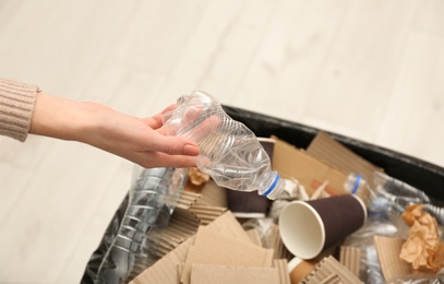 Photo of Young woman throwing plastic bottle in trash bin indoors, closeup. Waste recycling