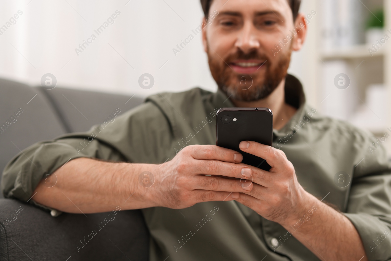 Photo of Smiling man using smartphone on sofa at home, selective focus