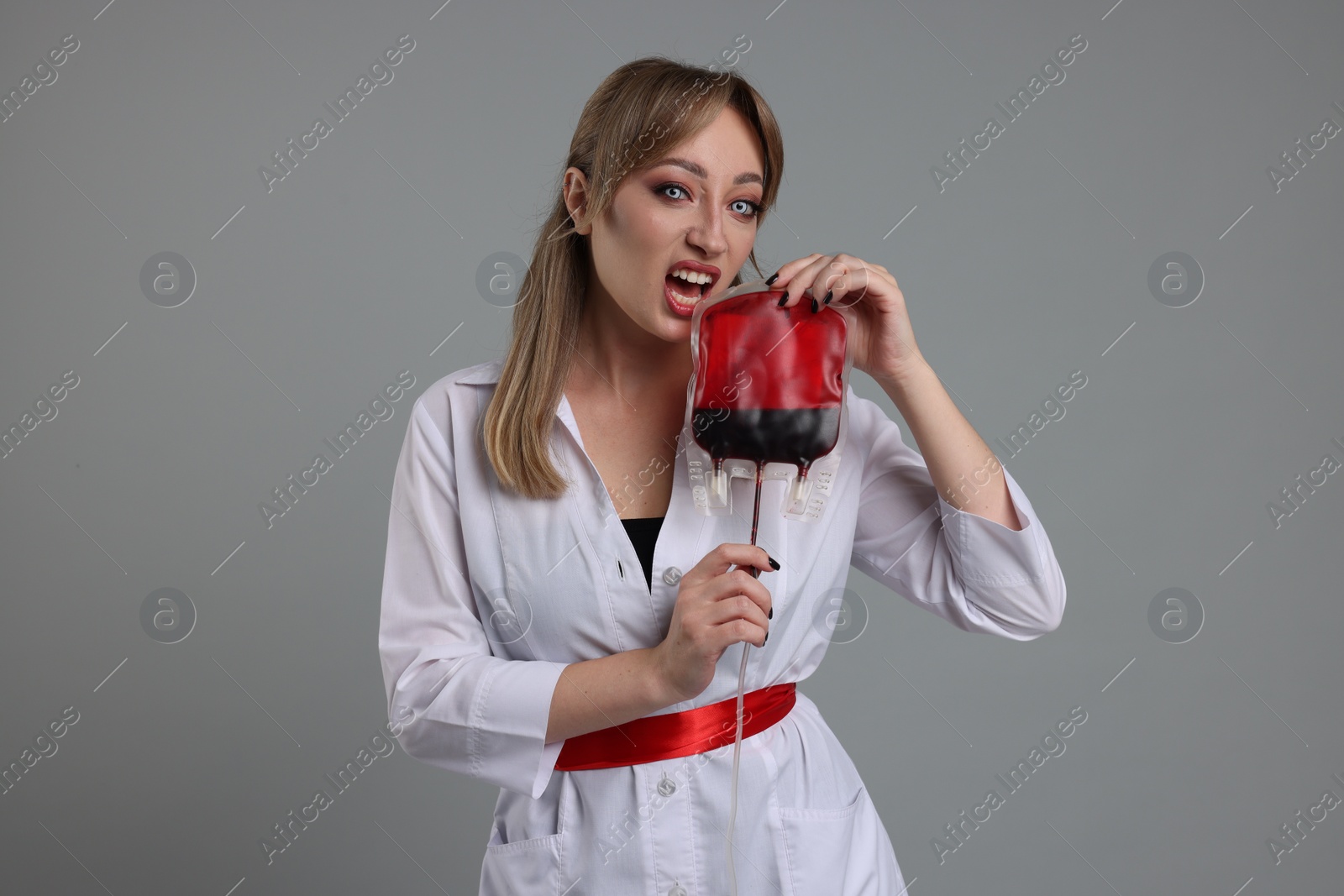 Photo of Woman in scary nurse costume with blood bag on light grey background. Halloween celebration