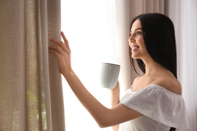 Woman with cup of coffee opening window curtain at home in morning