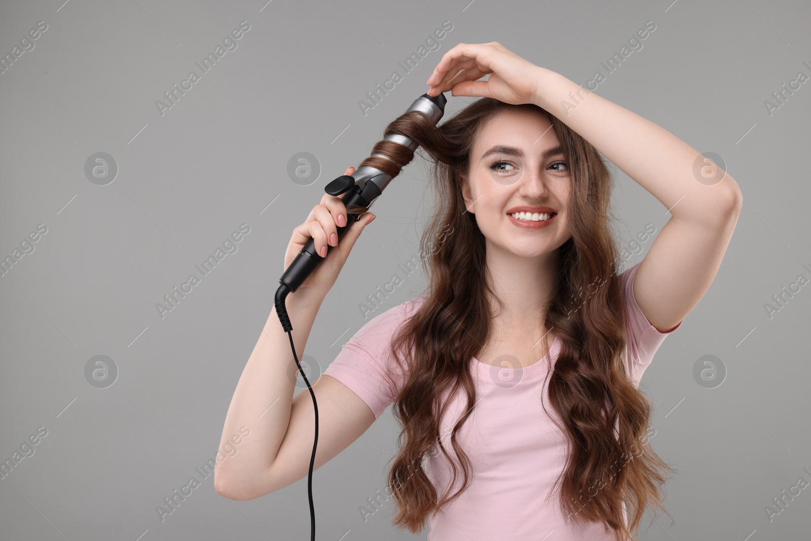 Photo of Beautiful young woman using curling hair iron on grey background, space for text