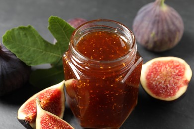 Glass jar with tasty sweet jam, green leaf and fresh figs on grey table, closeup
