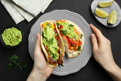 Photo of Woman holding taco with guacamole and vegetables at black table, top view