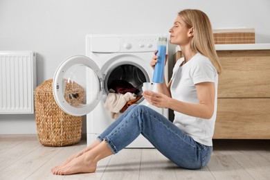 Photo of Woman sitting on floor near washing machine and smelling fabric softener in bathroom
