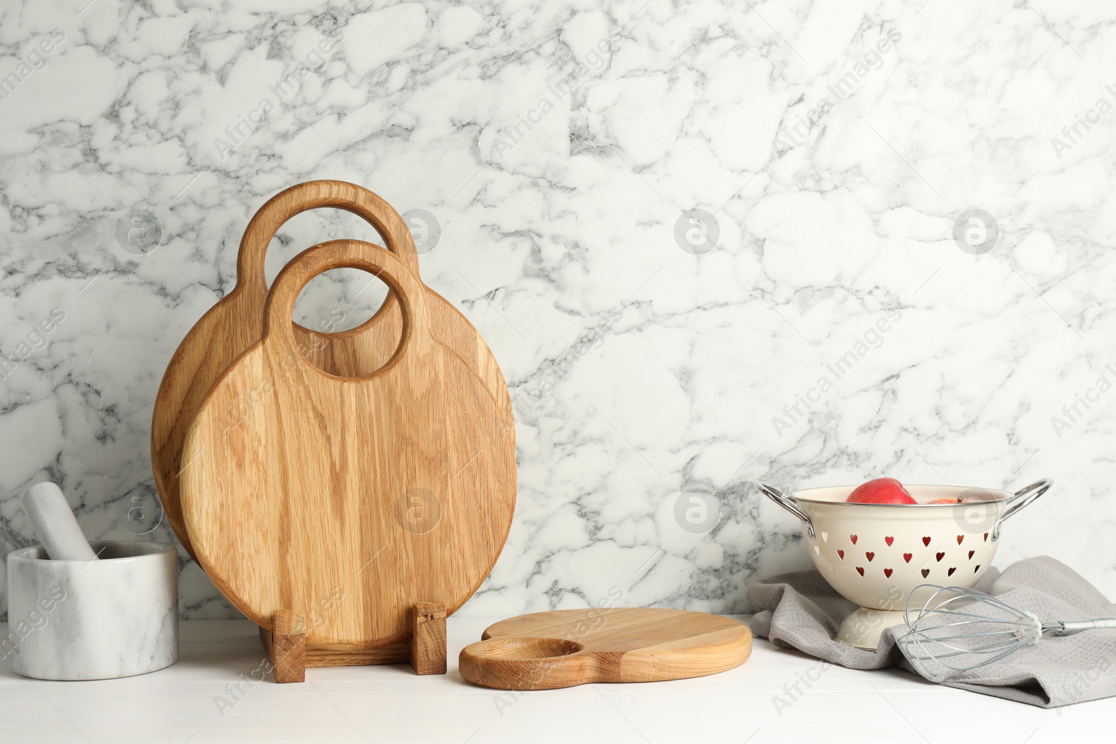 Photo of Wooden cutting boards, mortar with pestle and colander on white table near marble wall