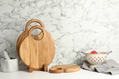 Photo of Wooden cutting boards, mortar with pestle and colander on white table near marble wall