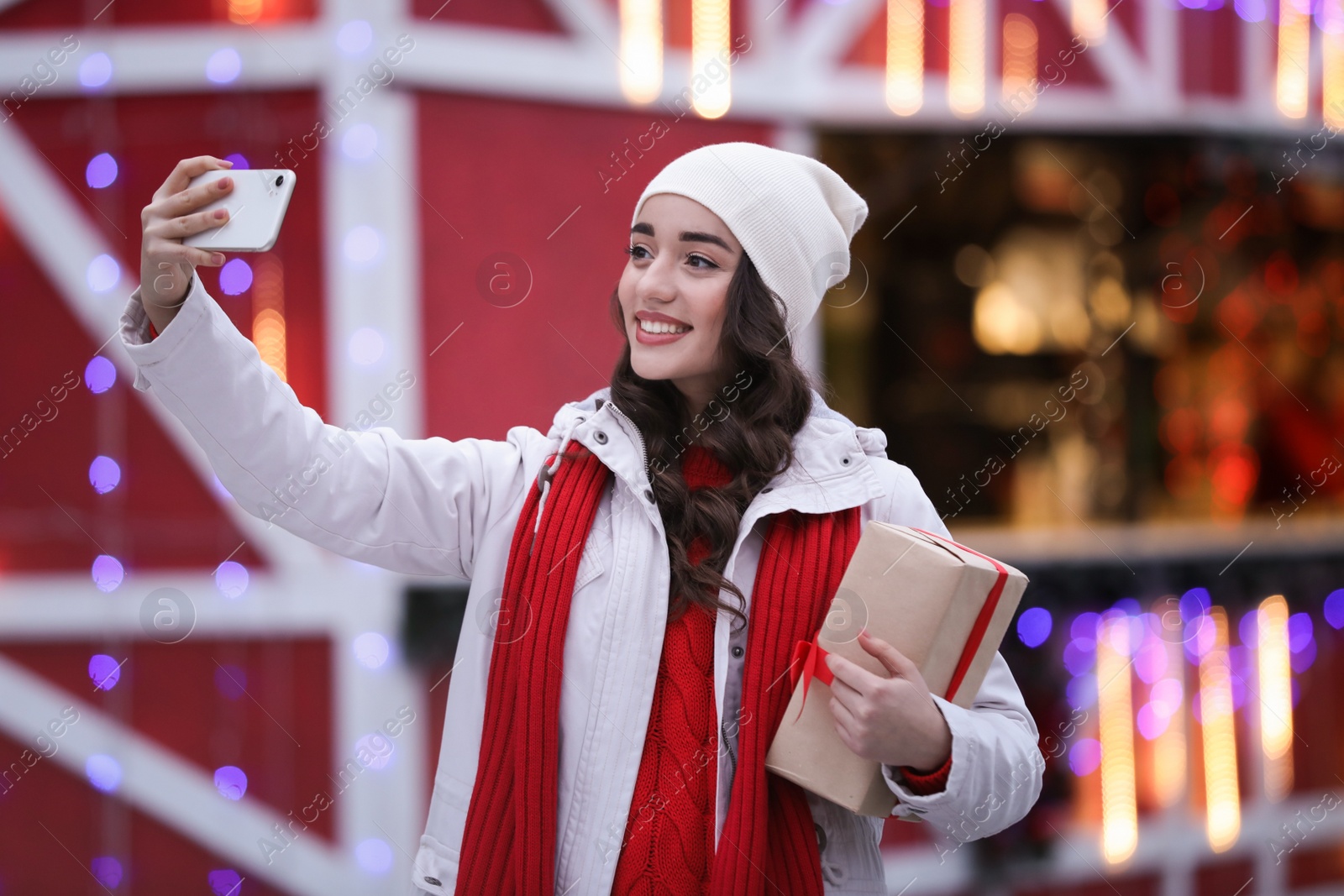 Photo of Happy young woman with gift box taking selfie at winter fair in evening. Christmas celebration