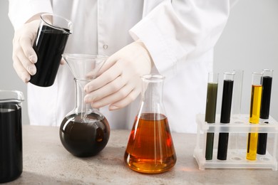 Photo of Woman pouring black crude oil from beaker into flask at grey table against light background, closeup