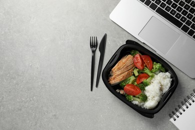 Photo of Container of tasty food, laptop, cutlery and notebook on light grey table, flat lay with space for text. Business lunch