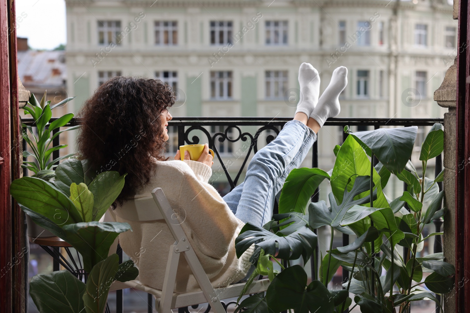 Photo of Young woman with cup of tea relaxing in chair surrounded by green houseplants on balcony