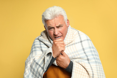 Senior man wrapped in blanket coughing on yellow background. Cold symptoms