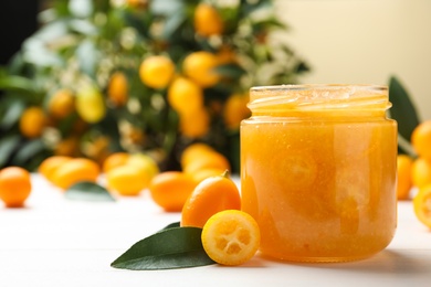 Photo of Delicious kumquat jam in jar and fresh fruits on white wooden table, space for text
