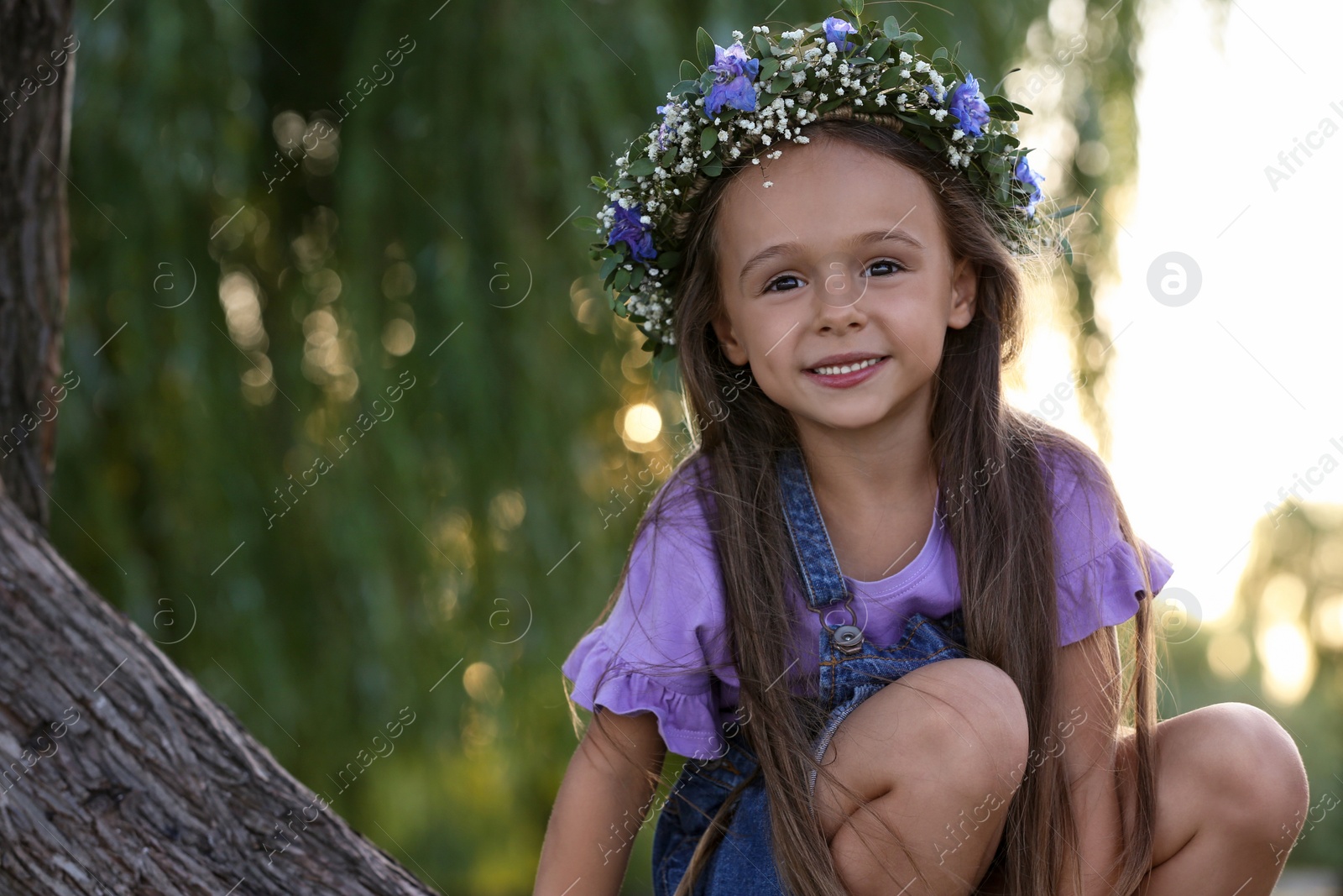 Photo of Cute little girl wearing flower wreath on tree outdoors. Child spending time in nature