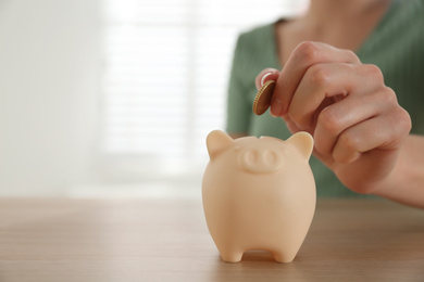 Photo of Woman putting money into piggy bank at wooden table indoors, closeup. Space for text