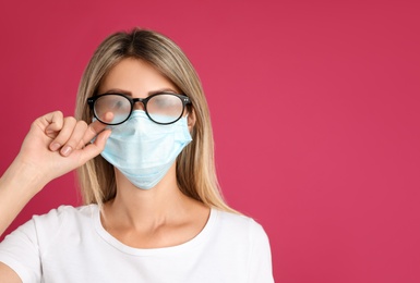 Photo of Woman wiping foggy glasses caused by wearing disposable mask on pink background, space for text. Protective measure during coronavirus pandemic