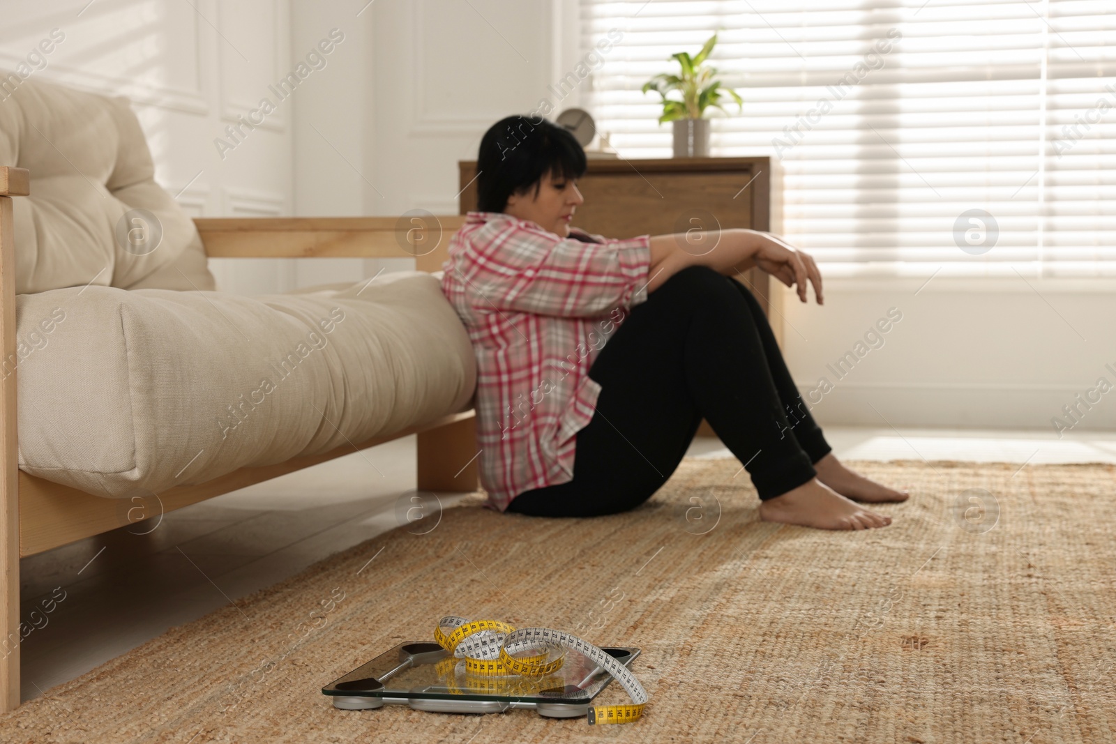 Photo of Depressed overweight woman sitting near sofa at home, focus on scales with measuring tape
