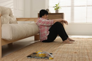 Depressed overweight woman sitting near sofa at home, focus on scales with measuring tape