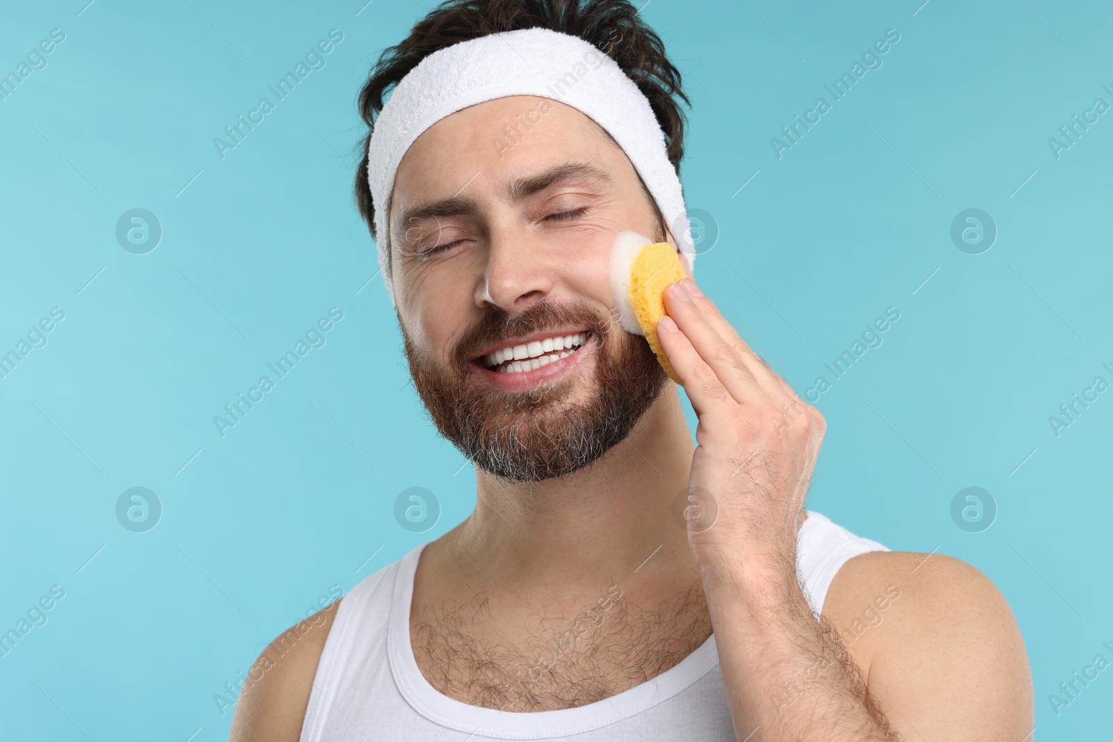 Photo of Man with headband washing his face using sponge on light blue background