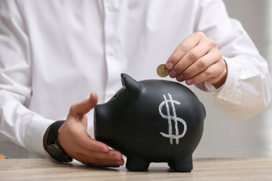 Photo of Man putting coin into piggy bank with dollar sign at wooden table, closeup