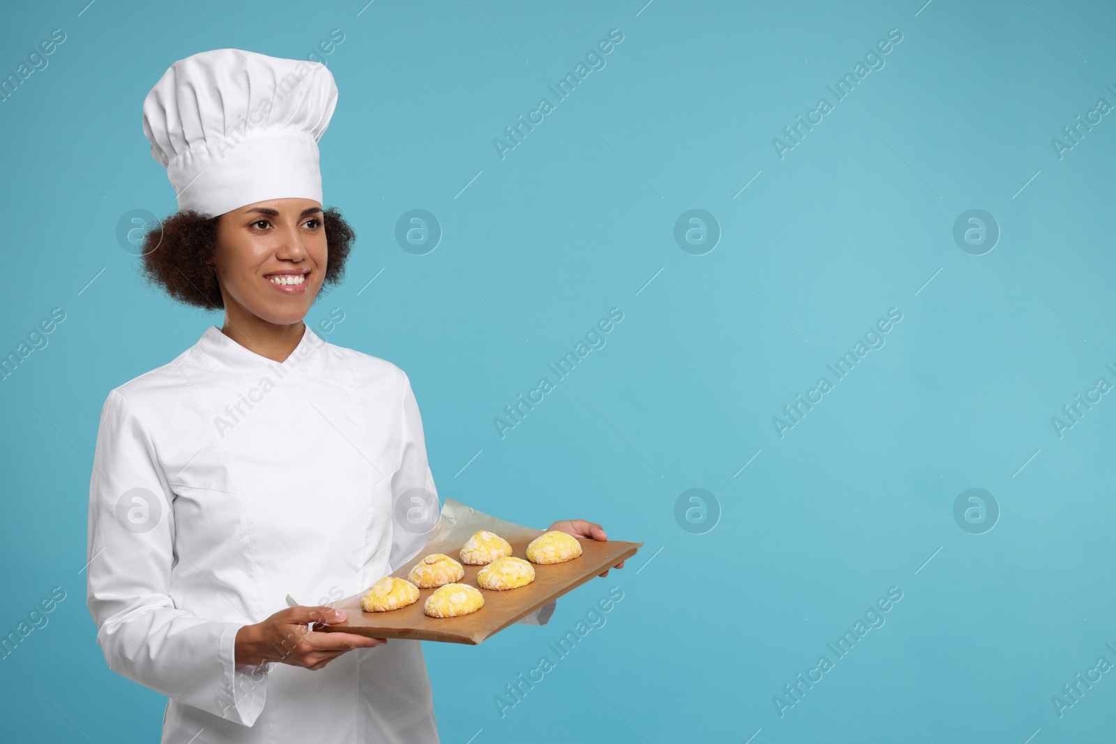 Photo of Happy female chef in uniform holding board with cookies on light blue background. Space for text