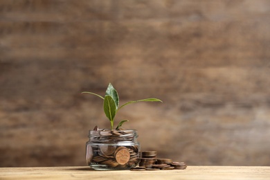 Photo of Glass jar and coins with young green plant on wooden table, space for text
