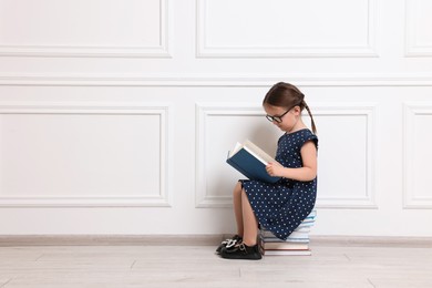 Cute little girl reading on stack of books near white wall. Space for text