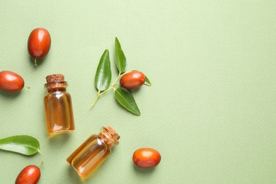 Glass bottles with jojoba oil and seeds on green background, flat lay. Space for text