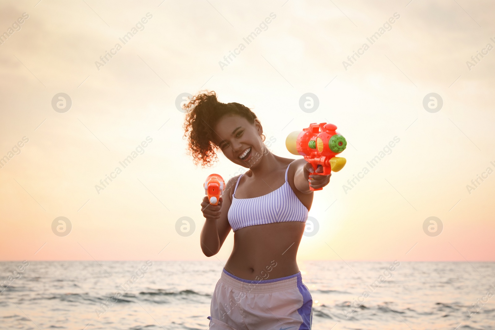 Photo of African American woman with water guns having fun on beach at sunset