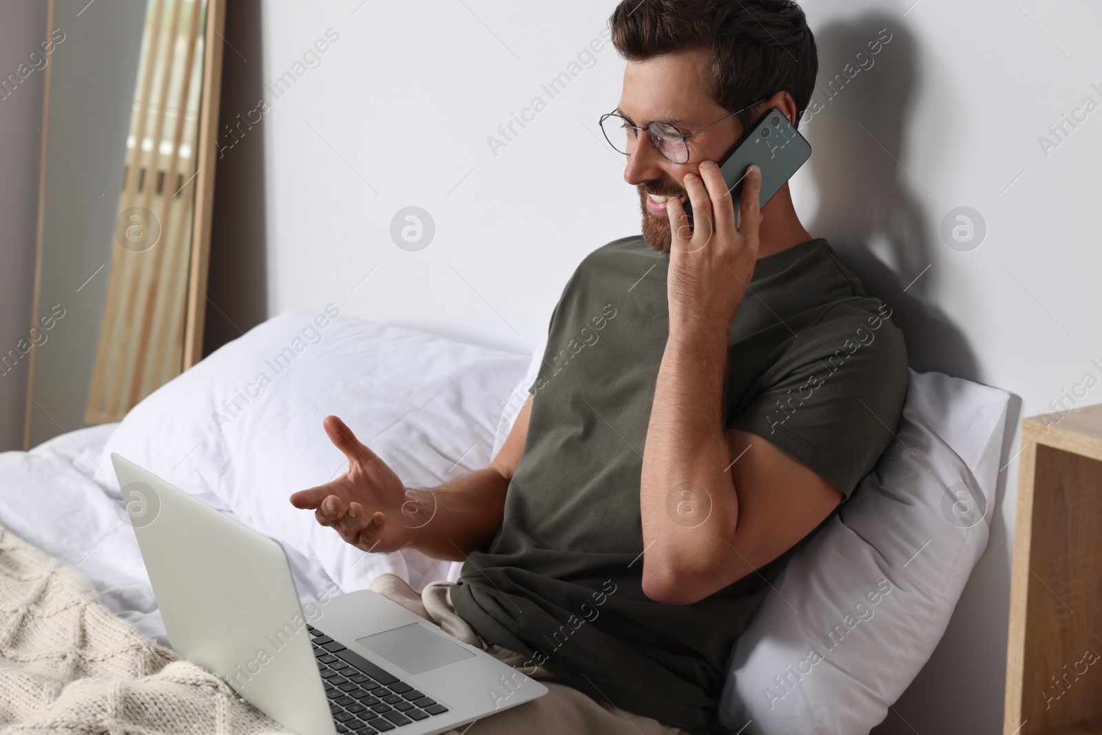 Photo of Handsome man talking on smartphone while working with laptop in bed at home