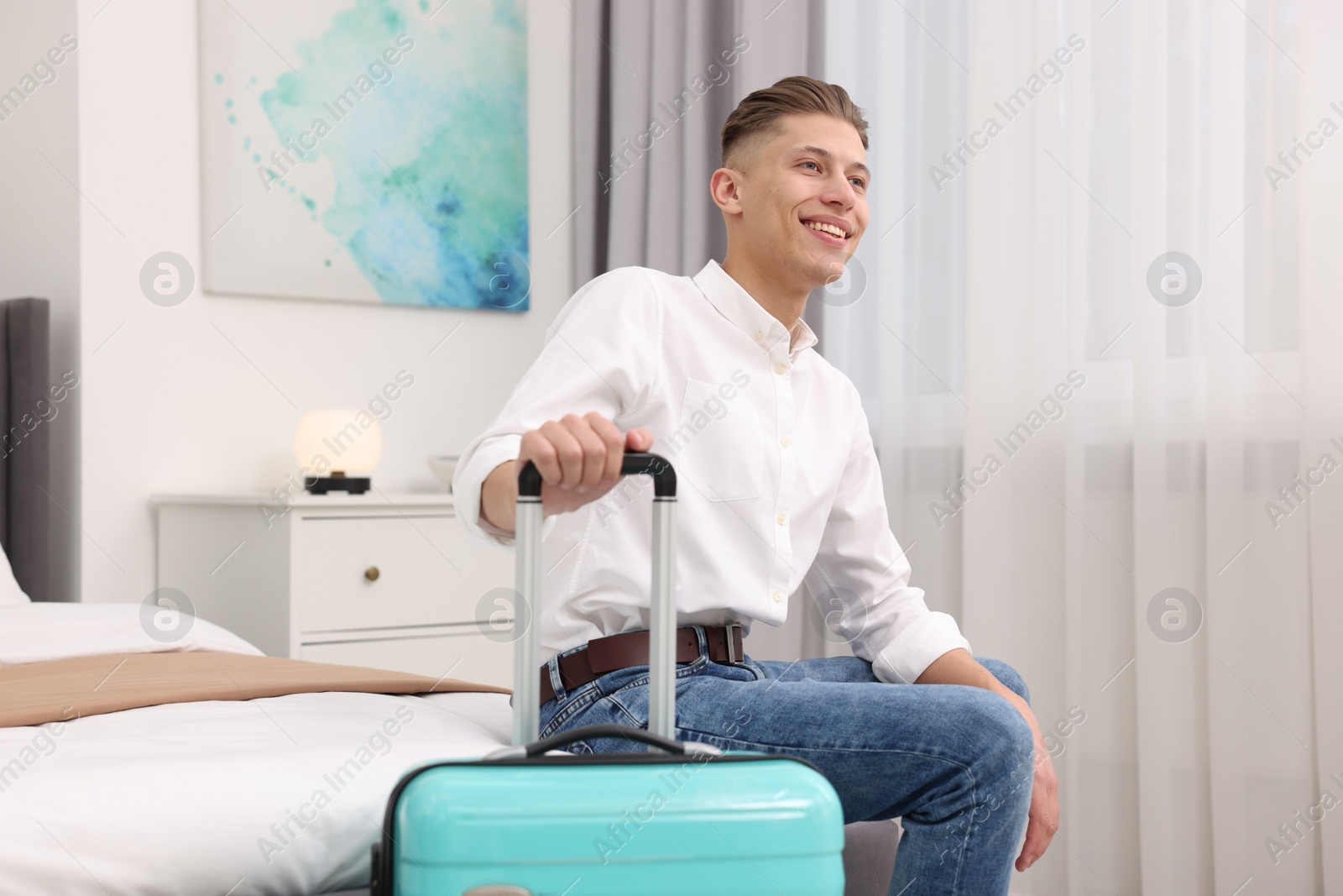 Photo of Smiling guest with suitcase relaxing on bed in stylish hotel room