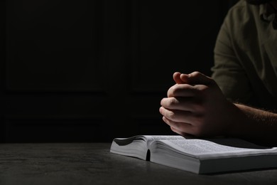Photo of Religion. Christian man praying over Bible at table against black background, closeup. Space for text