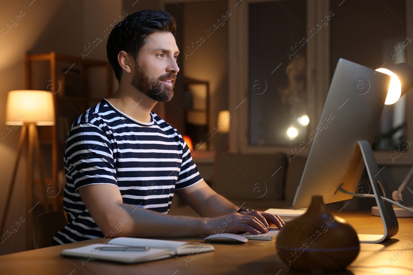 Photo of Home workplace. Man working with computer at wooden desk in room at night