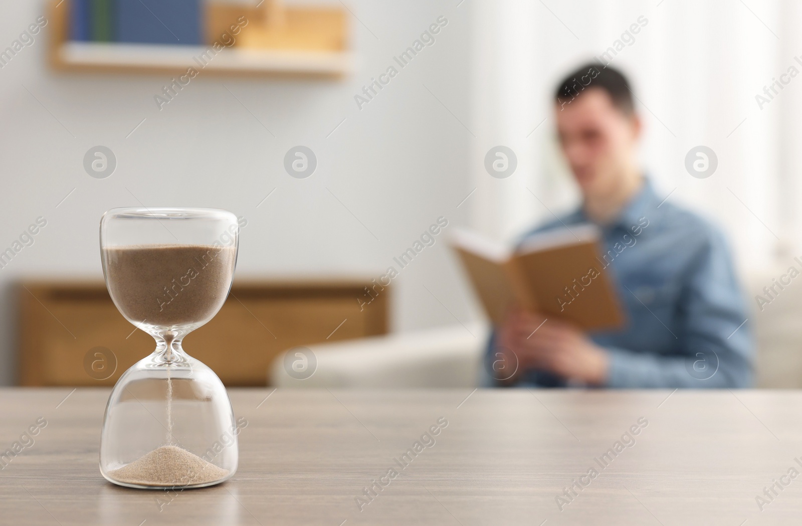 Photo of Hourglass with flowing sand on desk. Man reading book in room, selective focus