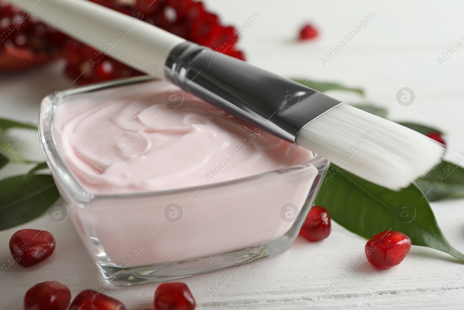 Photo of Glass bowl with natural facial mask, pomegranate seeds and brush on white wooden table, closeup