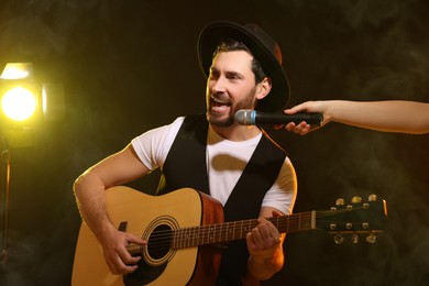 Image of Handsome man with acoustic guitar singing while woman holding microphone on stage in color lighted smoke