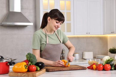 Happy young housewife cutting bell pepper at white marble table in kitchen