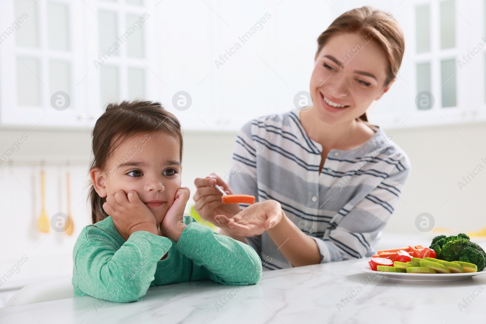 Photo of Mother feeding her daughter in kitchen. Little girl refusing to eat vegetables