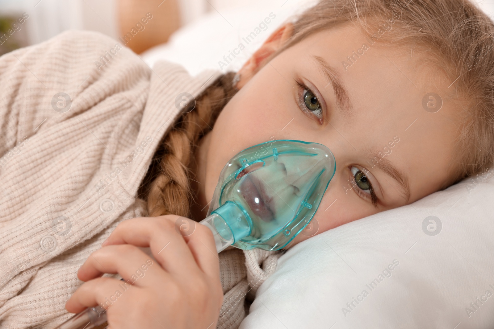 Photo of Little girl using nebulizer for inhalation on bed indoors, closeup