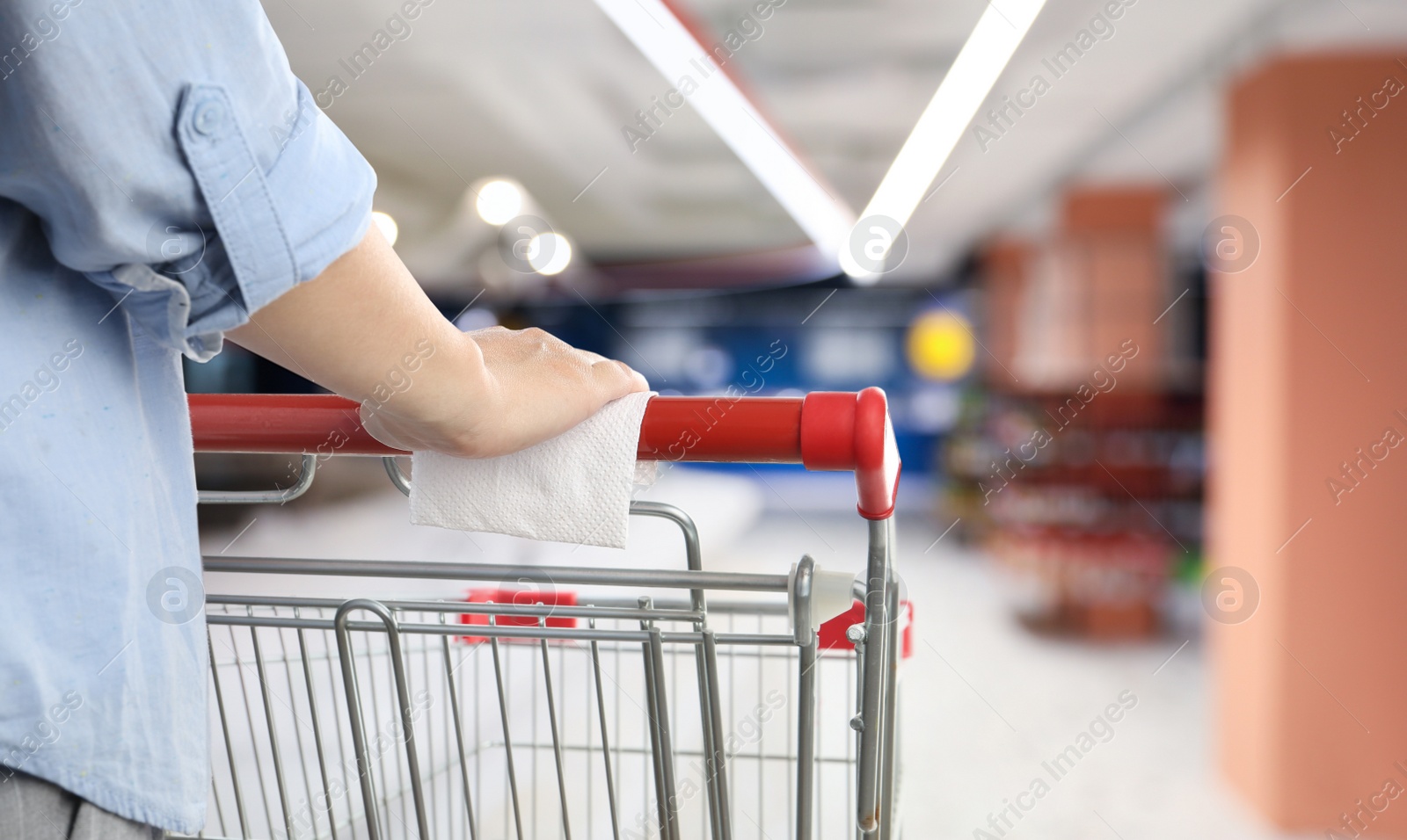 Image of Woman holding shopping cart handle with tissue paper at supermarket, closeup. Preventive measure in public places during coronavirus outbreak