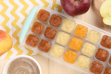 Photo of Different purees in ice cube tray and fresh apple fruits on wooden table, flat lay. Ready for freezing