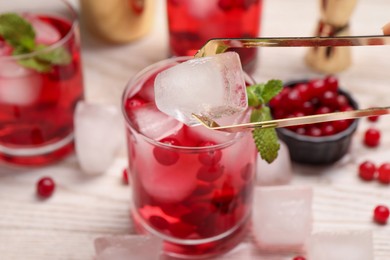 Photo of Putting ice cube into glass with tasty cranberry cocktail at light table, closeup