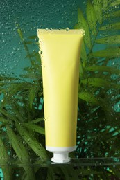 Tube with moisturizing cream and palm leaves on green background, view through wet glass