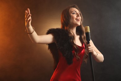 Photo of Beautiful young woman in stylish dress with microphone singing on dark background in color lights and smoke