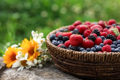 Photo of Wicker bowl with different fresh ripe berries and beautiful flowers on wooden surface outdoors, closeup